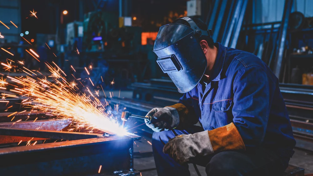 A welder working on a joint of two steel bars.